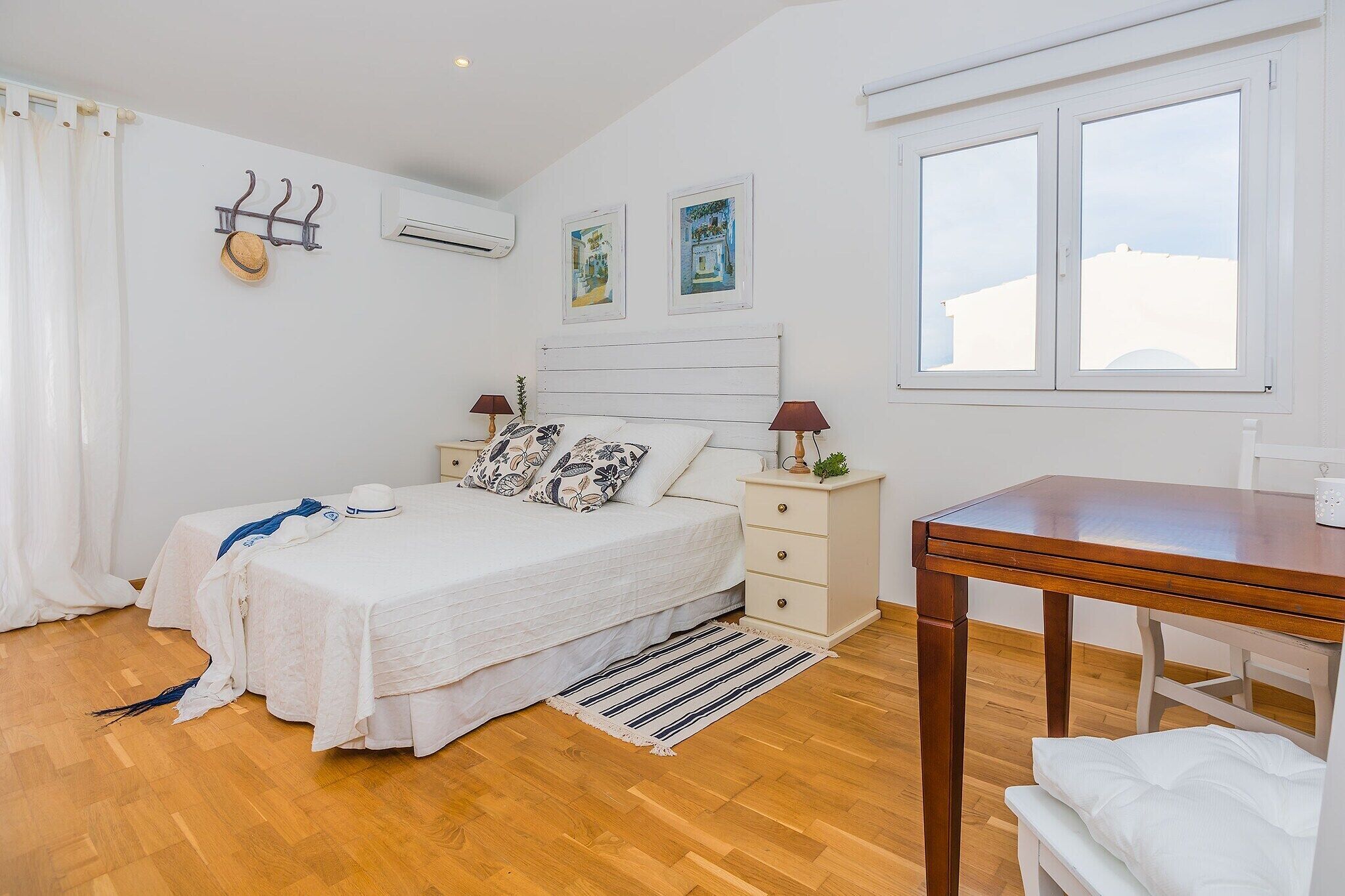 A bedroom with a bed, bedside tables, a desk, a window, and artwork on the wall.