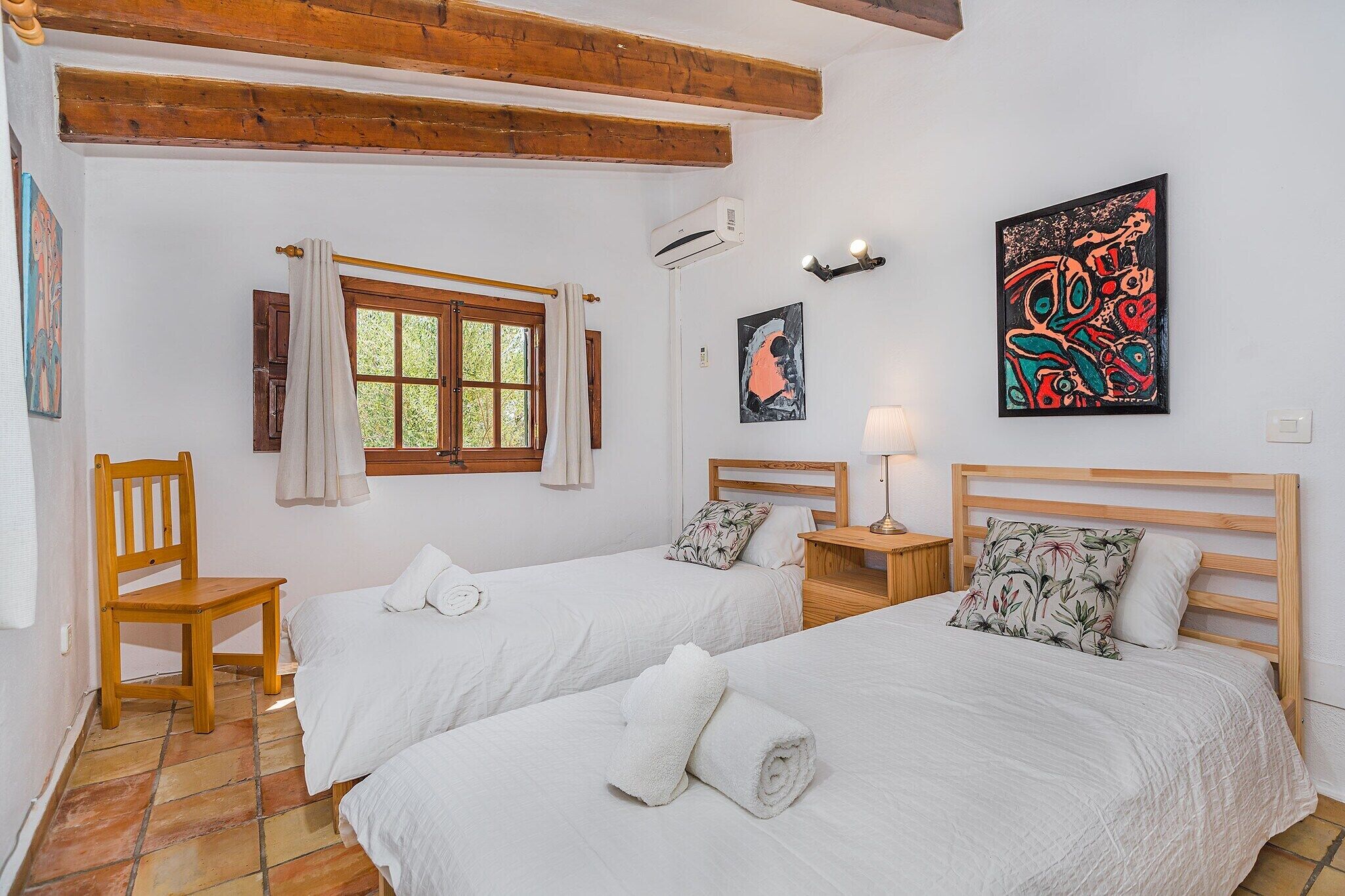 A room with two beds, a window, a chair, a lamp, and artwork on the walls.