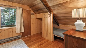 Un interno in legno con soffitto spiovente, una finestra con tende, un letto, un comodino con una lampada e un termosifone.
