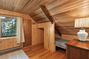 Un intérieur en bois avec un plafond en pente, une fenêtre avec des rideaux, un lit, une table de chevet avec une lampe et un radiateur.