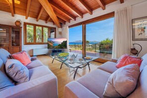 A modern living room with a glass coffee table, sofas, and a large window offering a scenic view. - 5-Bed Villa, Two Pools, Puerto Pollença (Pollensa / Pollença)