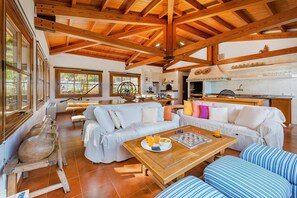 A spacious living area with a wooden ceiling, a sofa set, a coffee table with breakfast items, and a kitchen area with a stove and shelves. - 5-Bed Villa, Two Pools, Puerto Pollença (Pollensa / Pollença)