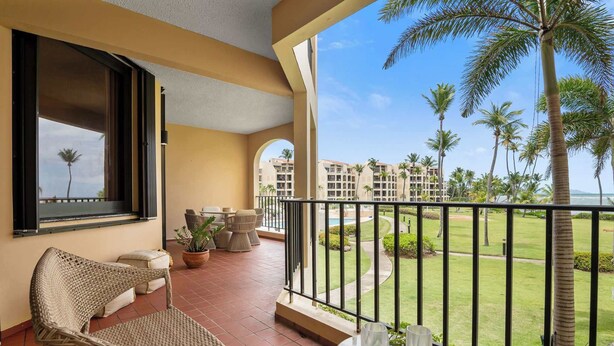 Een balkon met rieten meubilair, een plant in pot en uitzicht op een resort met palmbomen en een zwembad.