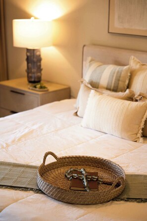 A neatly made bed with a woven basket containing a bottle and a card, a bedside lamp, and a nightstand with a small plant. - Boho 2BR Retreat - Ocean View Terrace & Pool (Palmas del Mar)