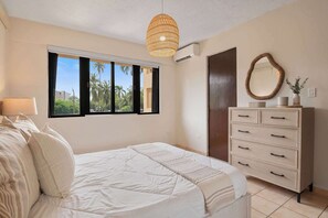 A bedroom with a bed, a dresser, a window with a view of palm trees, and a woven light fixture. - Boho 2BR Retreat - Ocean View Terrace & Pool (Palmas del Mar)