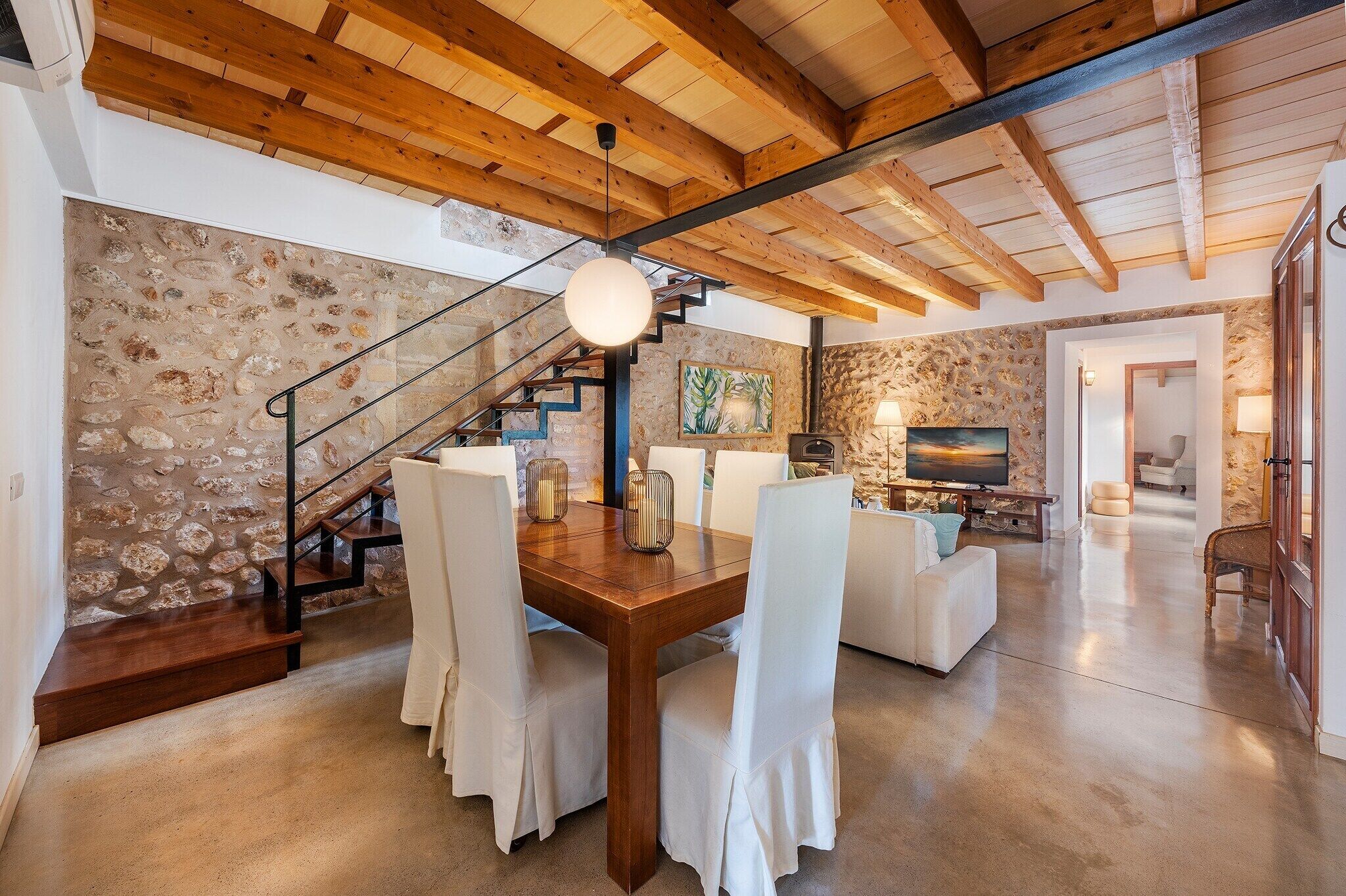 A spacious living area with a stone wall, wooden ceiling beams, a dining table with chairs, and a staircase.