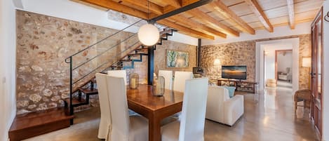 Una zona d'estar àmplia amb una paret de pedra, bigues de fusta al sostre, taula de dinar amb cadires i una escala.