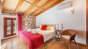 A bedroom with a wooden ceiling, stone wall, a bed with white bedding and red accents, a wicker chair, and a small table. - Villa near Pollença Town Centre (Pollensa / Pollença)