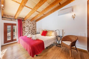 A bedroom with a wooden ceiling, stone wall, a bed with white bedding and red accents, a wicker chair, and a small table. - Villa near Pollença Town Centre (Pollensa / Pollença)