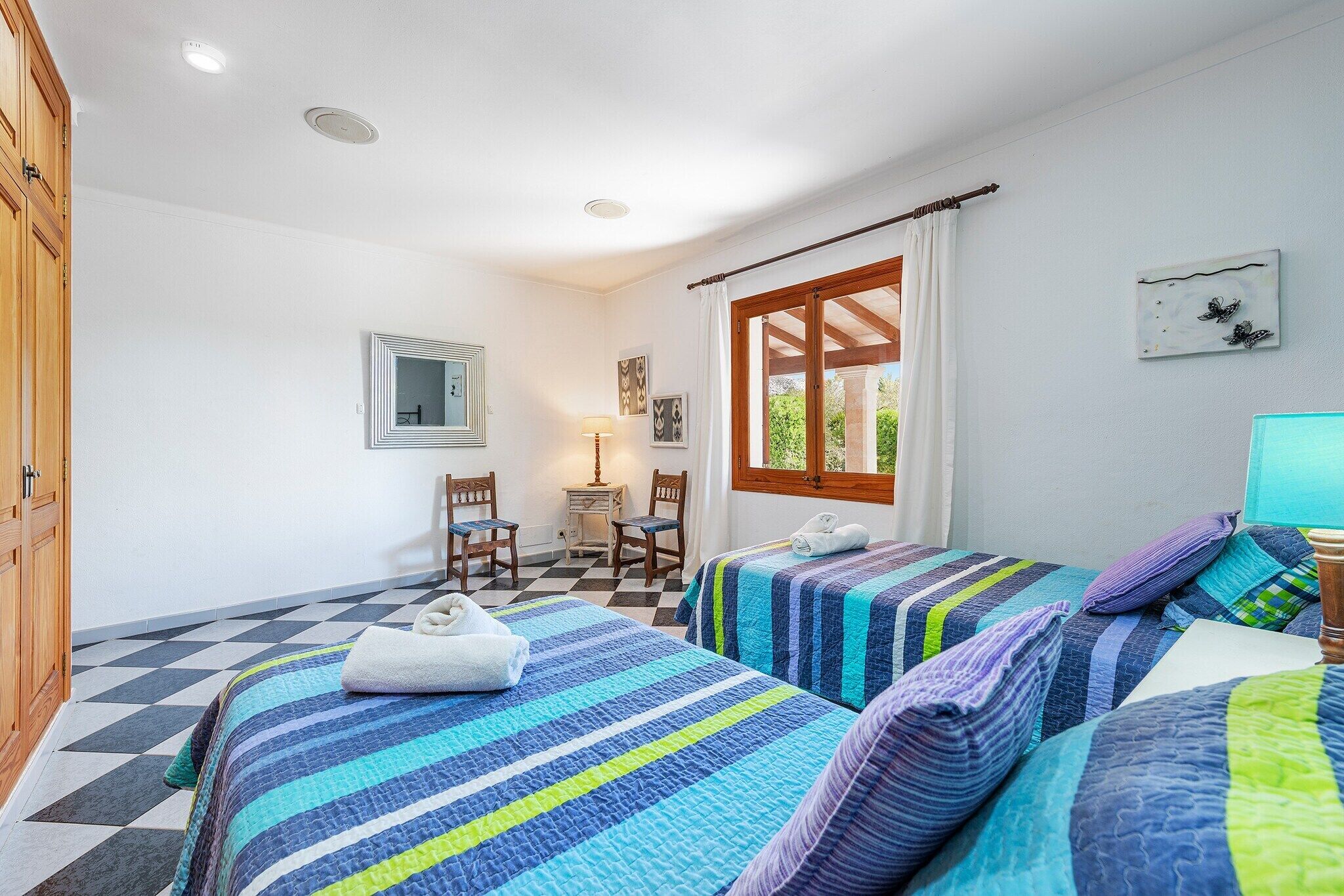 A bedroom with two beds, a wooden wardrobe, a desk with a chair, a window with curtains, and a wall-mounted mirror.