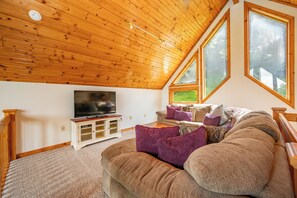A cozy living room with a wooden ceiling, a television on a cabinet, a sofa with cushions, and large windows. - New! Mountain Lake Area Getaway w/ Hot Tub! (Oakland)
