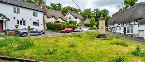 A rural street with traditional thatched-roof houses, a grassy area with a monument, and parked cars.