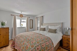 Ein Schlafzimmer mit einem Bett, einem hölzernen Schminktisch, einem Nachttisch, einem Fenster mit Vorhängen und einer Pendelleuchte.