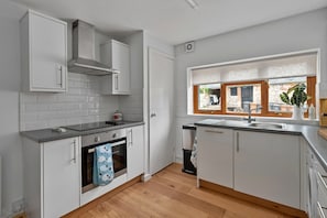A modern kitchen with white cabinetry, a built-in oven, a dishwasher, and a window with a view of the outside. - Romantic North Wales Meadow Cottage (Andover)