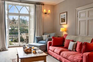Ein Wohnzimmer mit einem roten Sofa, einem blauen Sessel, einem Couchtisch mit Frühstücksgebäck und Blick durch ein großes Fenster.