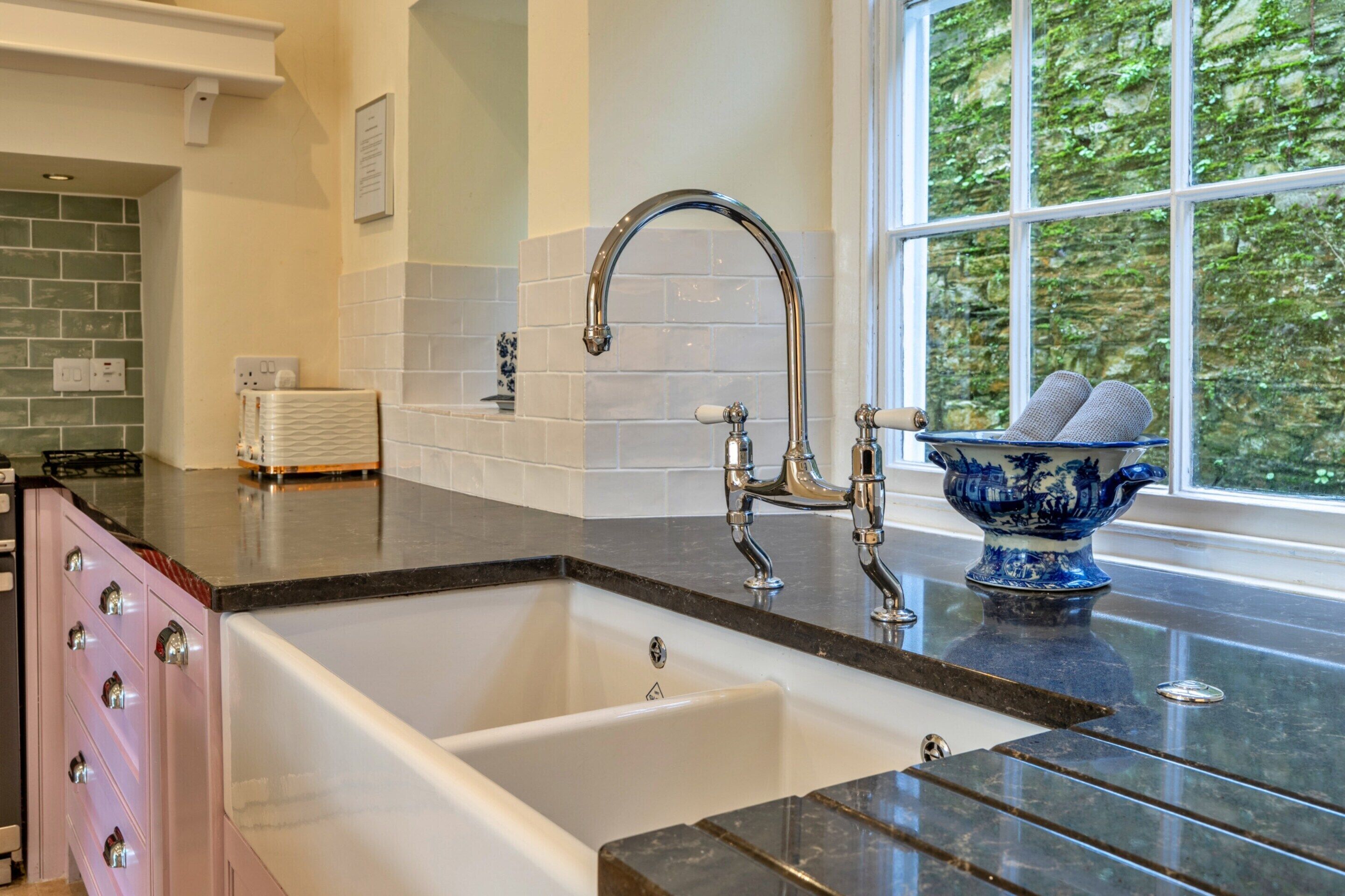 A kitchen with a white sink, a stainless steel faucet, a blue decorative bowl, and a window with a view of greenery.