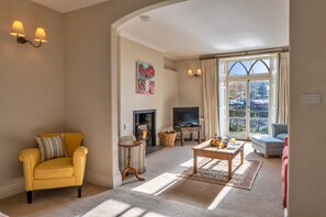 A living room with a yellow armchair, a fireplace, a television, and a view through a large window. - Pleasant Cottage in Banks of the River Fowey (Fowey)