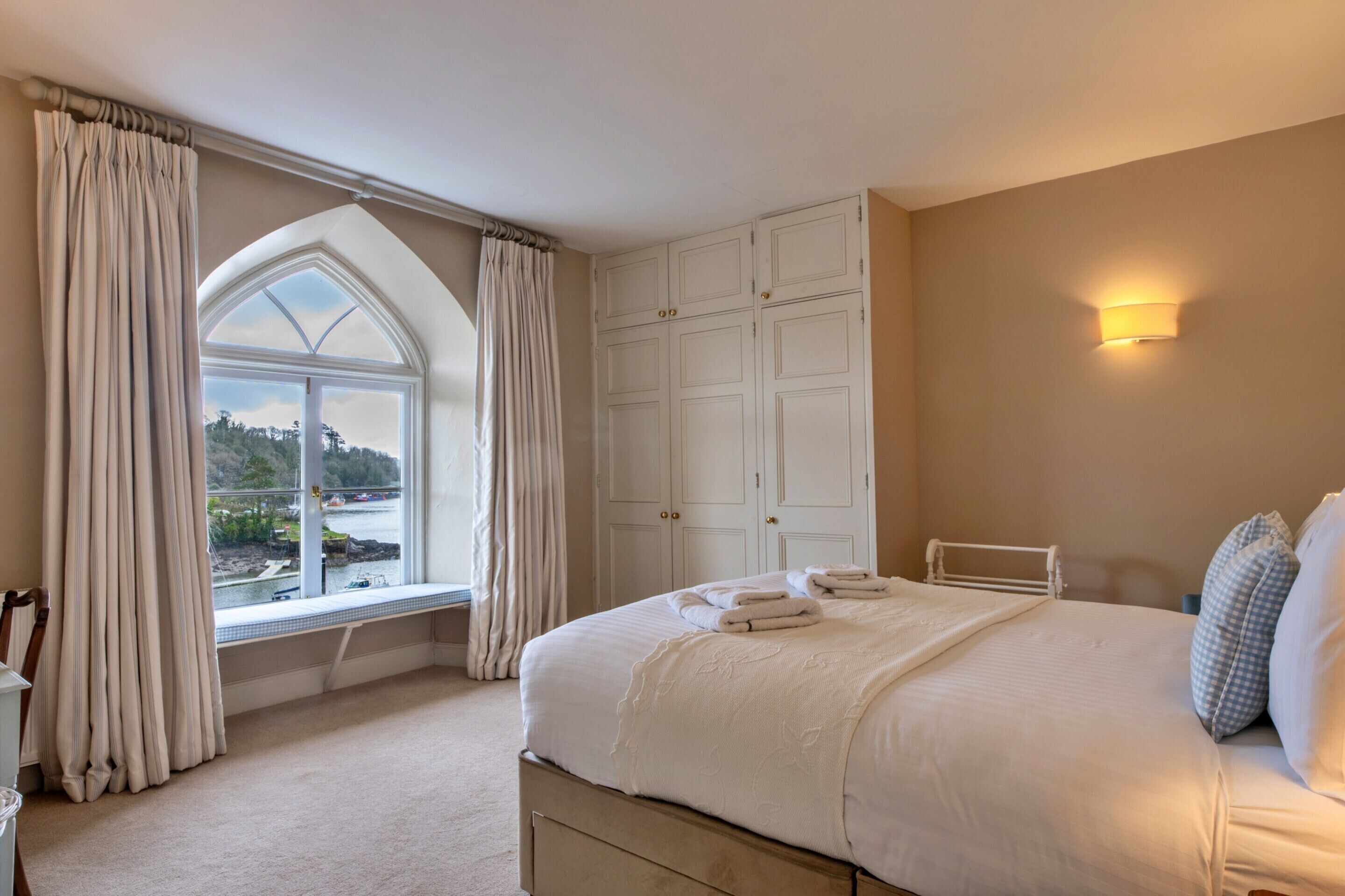 Een slaapkamer met een groot bed, een raam met gordijnen, een kledingkast en uitzicht op een waterpartij.