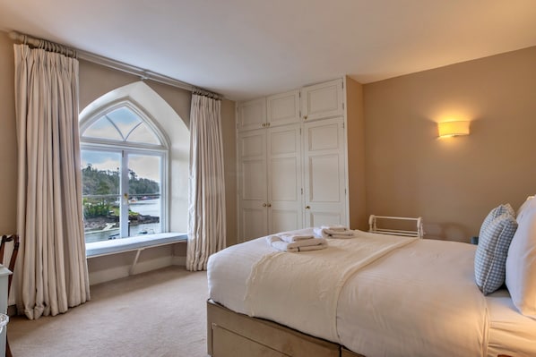 A bedroom with a large bed, a window with curtains, a wardrobe, and a view of a body of water. - Pleasant Cottage in Banks of the River Fowey (Fowey)