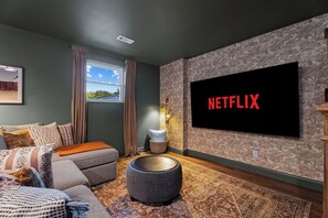 Ett modernt vardagsrum med en stor platt-TV som är monterad på väggen, en bekväm soffa, en rund pall och en mönstrad matta.