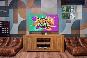 Ein Wohnzimmer mit Fernseher, der 'Party Fowl' zeigt, einer Holzkommode mit zwei Spielecontrollern und braunen Ledersofas.