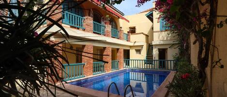 A courtyard with a swimming pool, a two-story building with balconies, and surrounding vegetation.