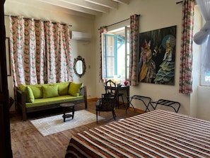 Una habitación con cama, sofá, mesa de comedor y sillas. Hay un cuadro en la pared y una ventana con cortinas.