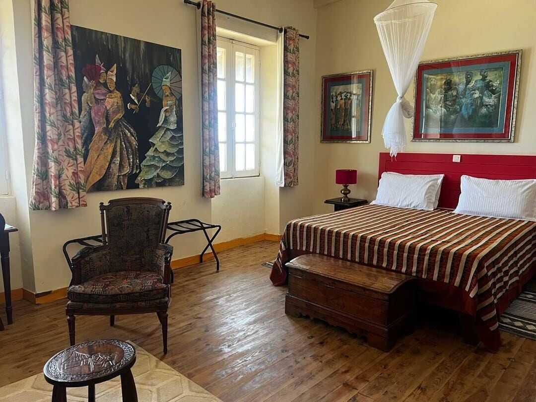 Ein Schlafzimmer mit einem Bett, einem Sessel, einem Nachttisch, einem Fenster mit Vorhängen und Bildern an den Wänden.