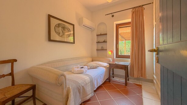 Ein Schlafzimmer mit Bett, Sessel, Nachttisch, Deckenventilator, Klimaanlage, Fenster mit Vorhängen und einer Wandregal.