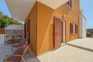 Ein modernes Gebäude mit flachem Dach, einer überdachten Außenbereich-Sitzgelegenheit mit Stühlen und einem Tisch sowie einer Tür mit Fenster.