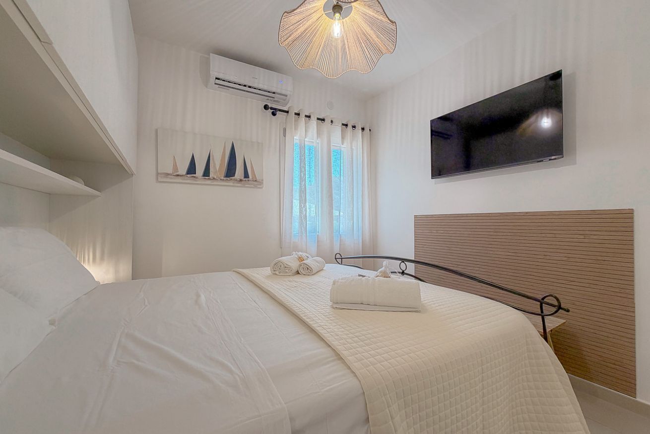 A hotel room with a bed, a flat-screen TV, an air conditioning unit, a window with curtains, and a wall-mounted artwork of sailboats.