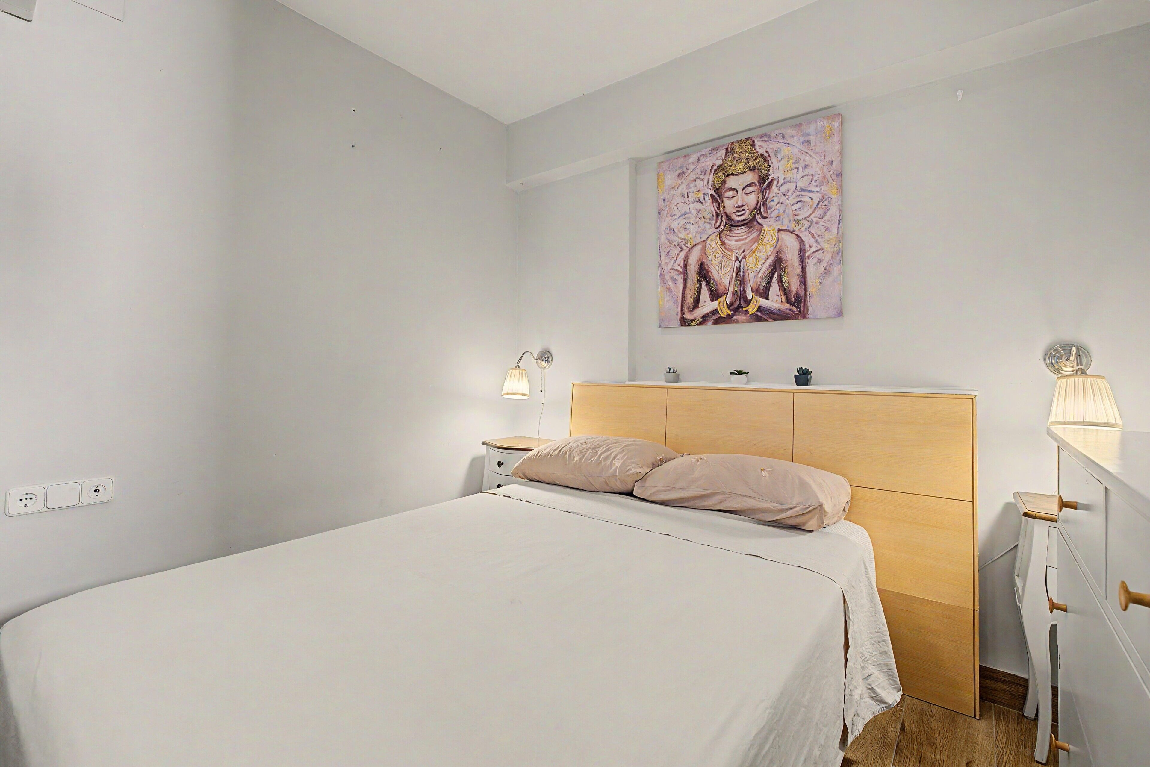 Een minimalistisch ingerichte slaapkamer met een bed, nachtlampjes, een houten hoofdbord en een schilderij aan de muur.