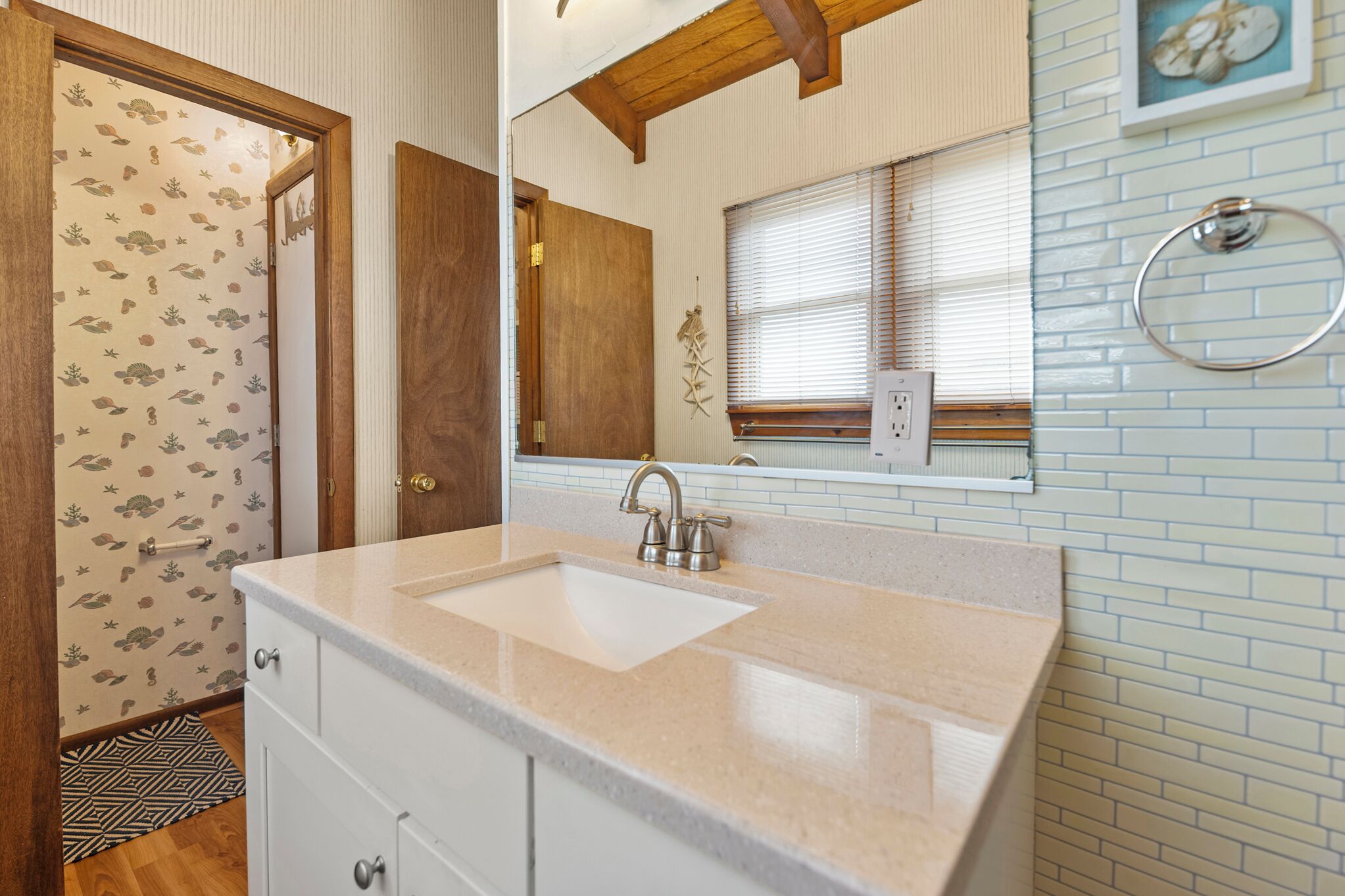 A bathroom with a sink, wooden door, patterned wallpaper, and a window with blinds.
