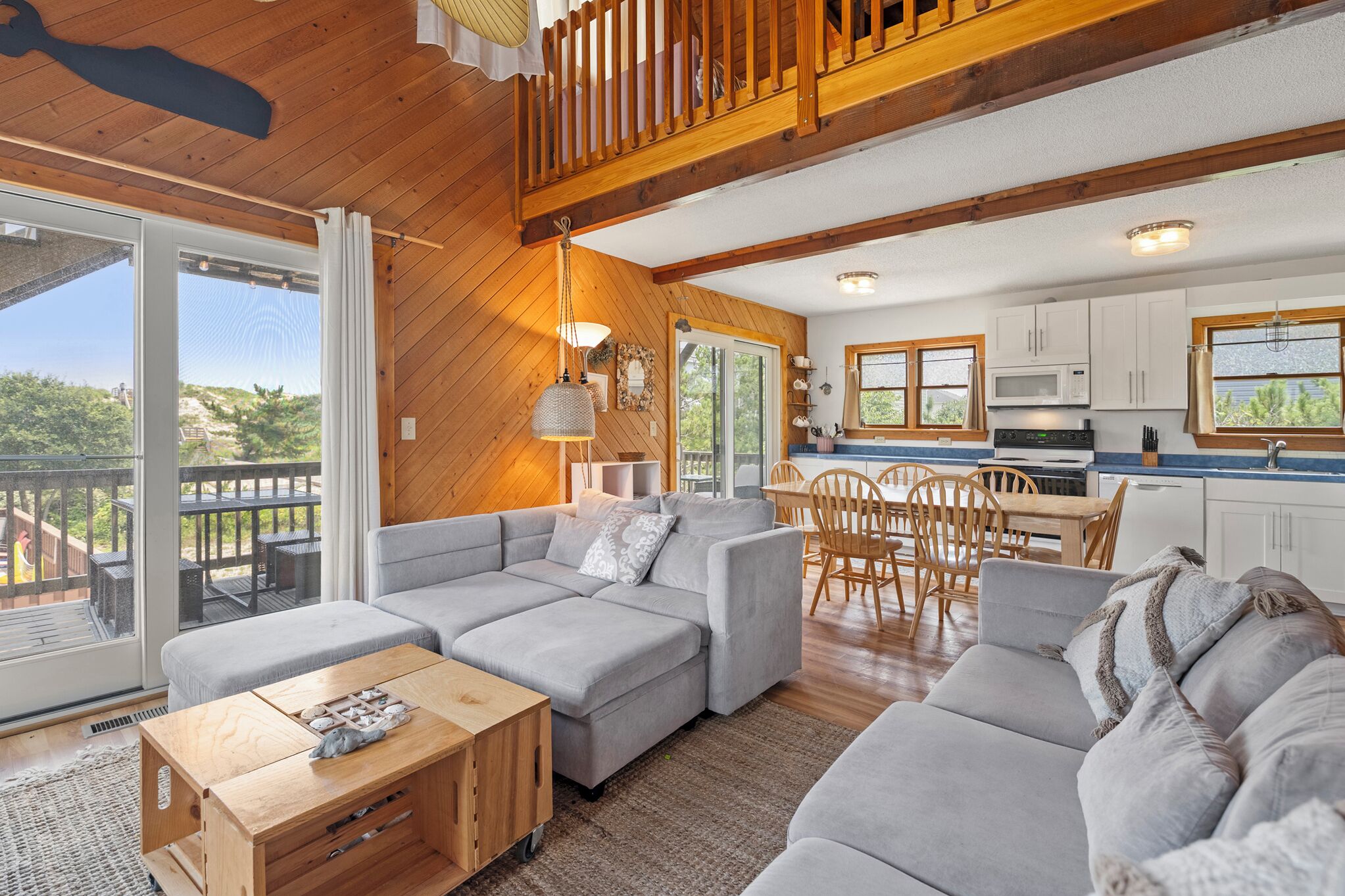 A spacious living area with a large grey sectional sofa, wooden flooring, and a balcony with a view.