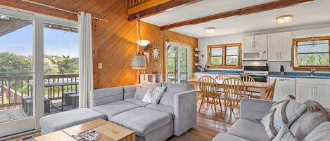 A spacious living area with a large grey sectional sofa, wooden flooring, and a balcony with a view.