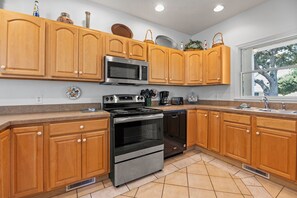 A kitchen with wooden cabinets, a stainless steel microwave, an oven, a dishwasher, and a window with a view of trees. - Calm C's: Dog Friendly | Hot Tub | Pool (Corolla)