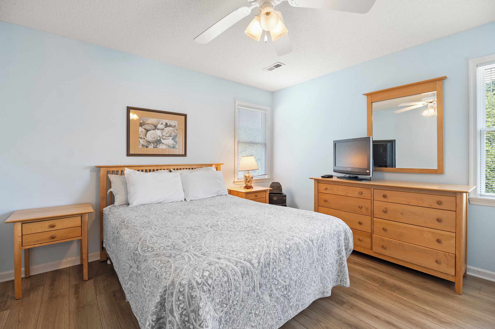 Une chambre à coucher comprenant un lit, une commode, une table de chevet, un ventilateur de plafond, une fenêtre avec des stores et un tableau encadré accroché au mur.