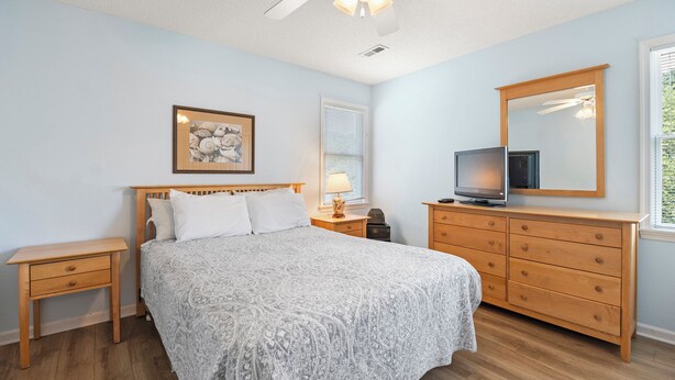 Um quarto com uma cama, cômoda, mesa de cabeceira, ventilador de teto, janela com persianas e uma moldura com um quadro na parede.
