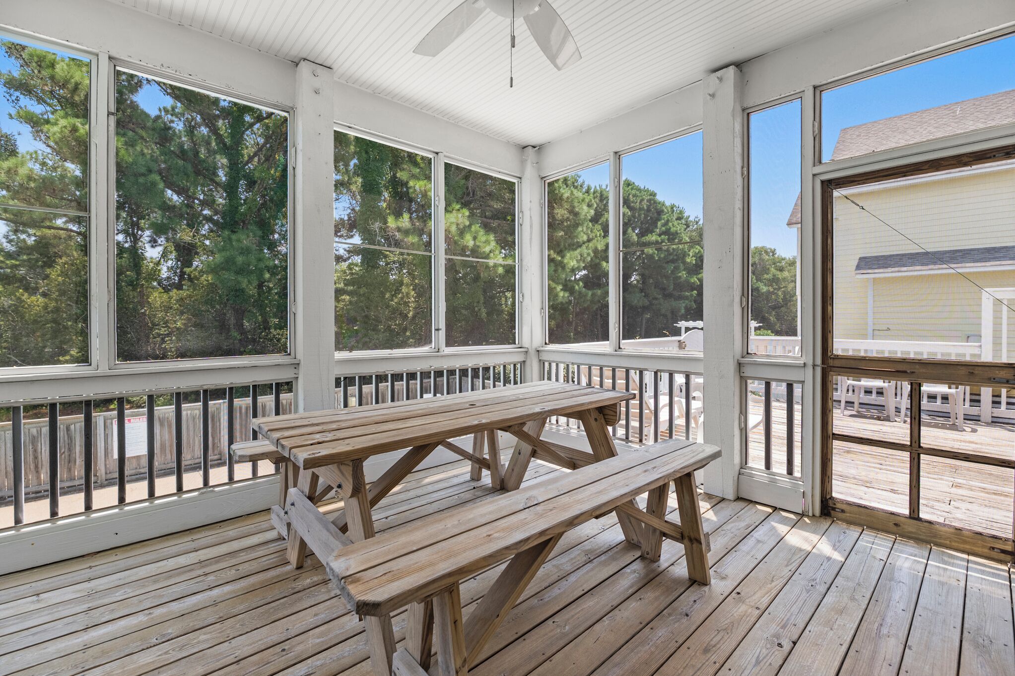 A covered outdoor area with a wooden picnic table and benches, surrounded by white railings and large windows.