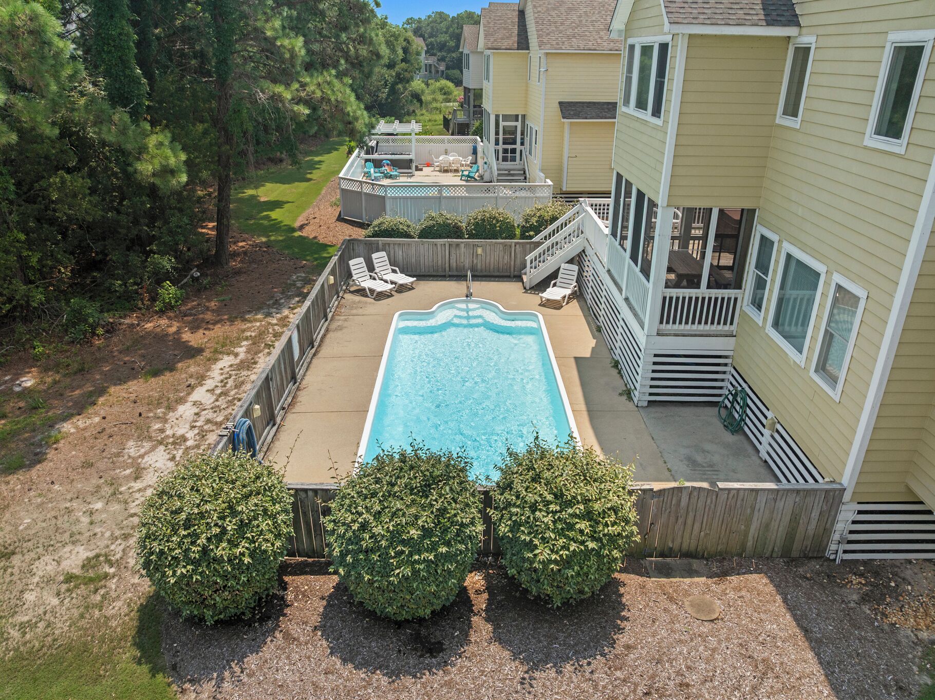 A rectangular swimming pool surrounded by a wooden deck with lounge chairs, a wooden fence, and a house in the background.