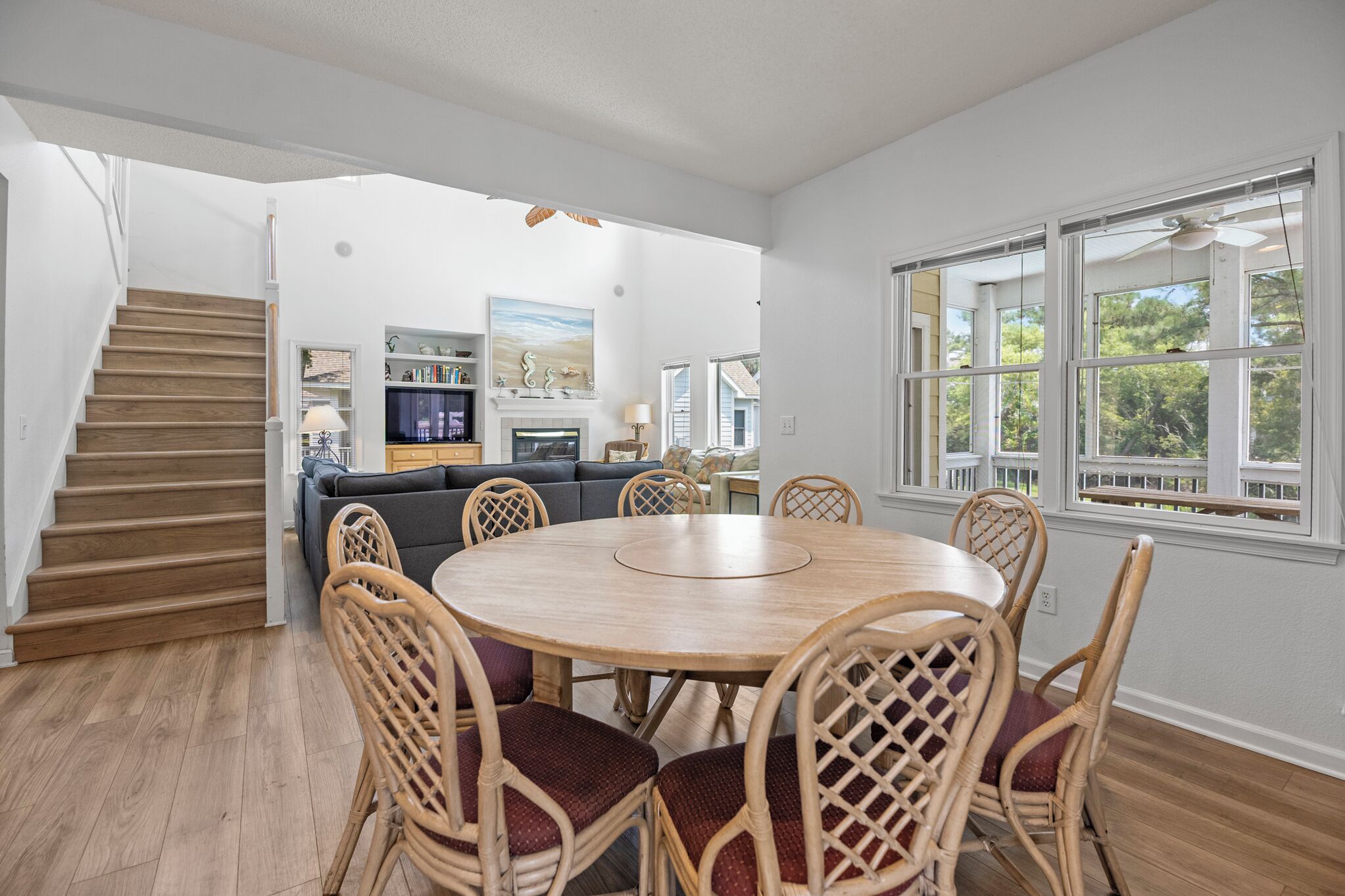 Een ruime woonkamer met een ronde houten eettafel omringd door rieten stoelen, een trap naar een hoger gelegen verdieping en een groot raam met uitzicht op bomen.