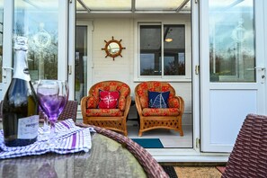 A patio area with wicker chairs, a table with a bottle and glass, and a door leading to a room with a ship's wheel decoration. - Historic Wiltshire Garden Cottage (Winchelsea)