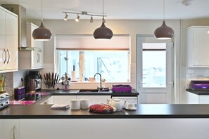 A modern kitchen with a central island, pendant lights, and a window with blinds. - Historic Wiltshire Garden Cottage (Winchelsea)