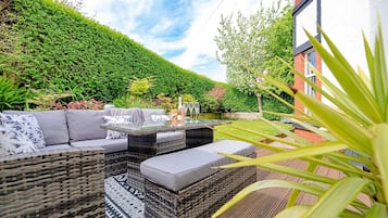 A patio with wicker furniture, a glass-top table, and a hedge in the background.