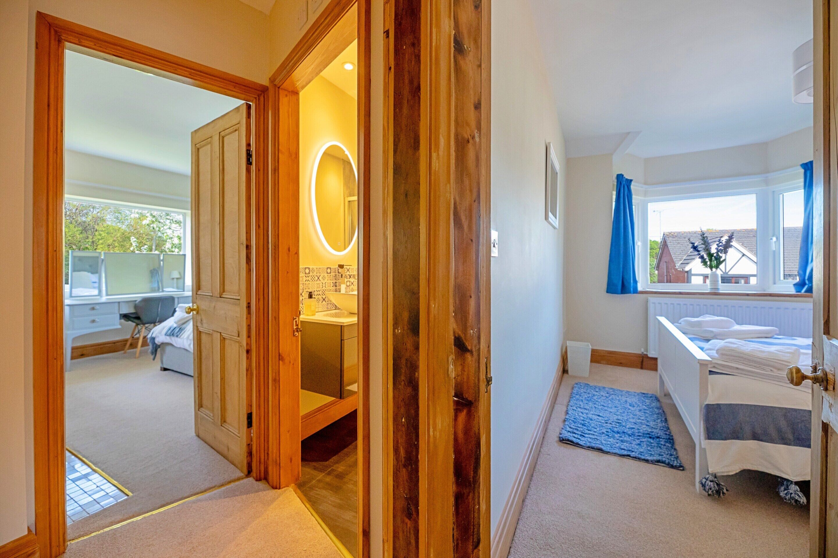 A hotel room with a wooden door, a bathroom with a mirror, a bed with a blue rug, and a window with curtains.