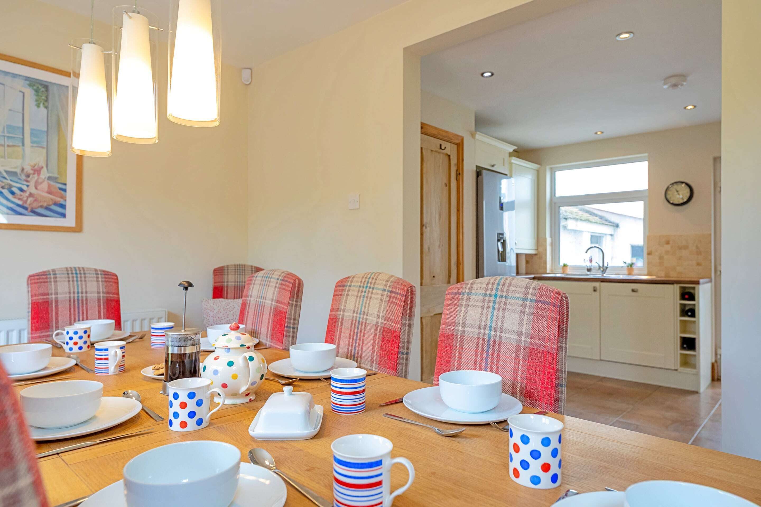 A dining area with a wooden table set for a meal, featuring a coffee pot, teapot, and various mugs and plates.