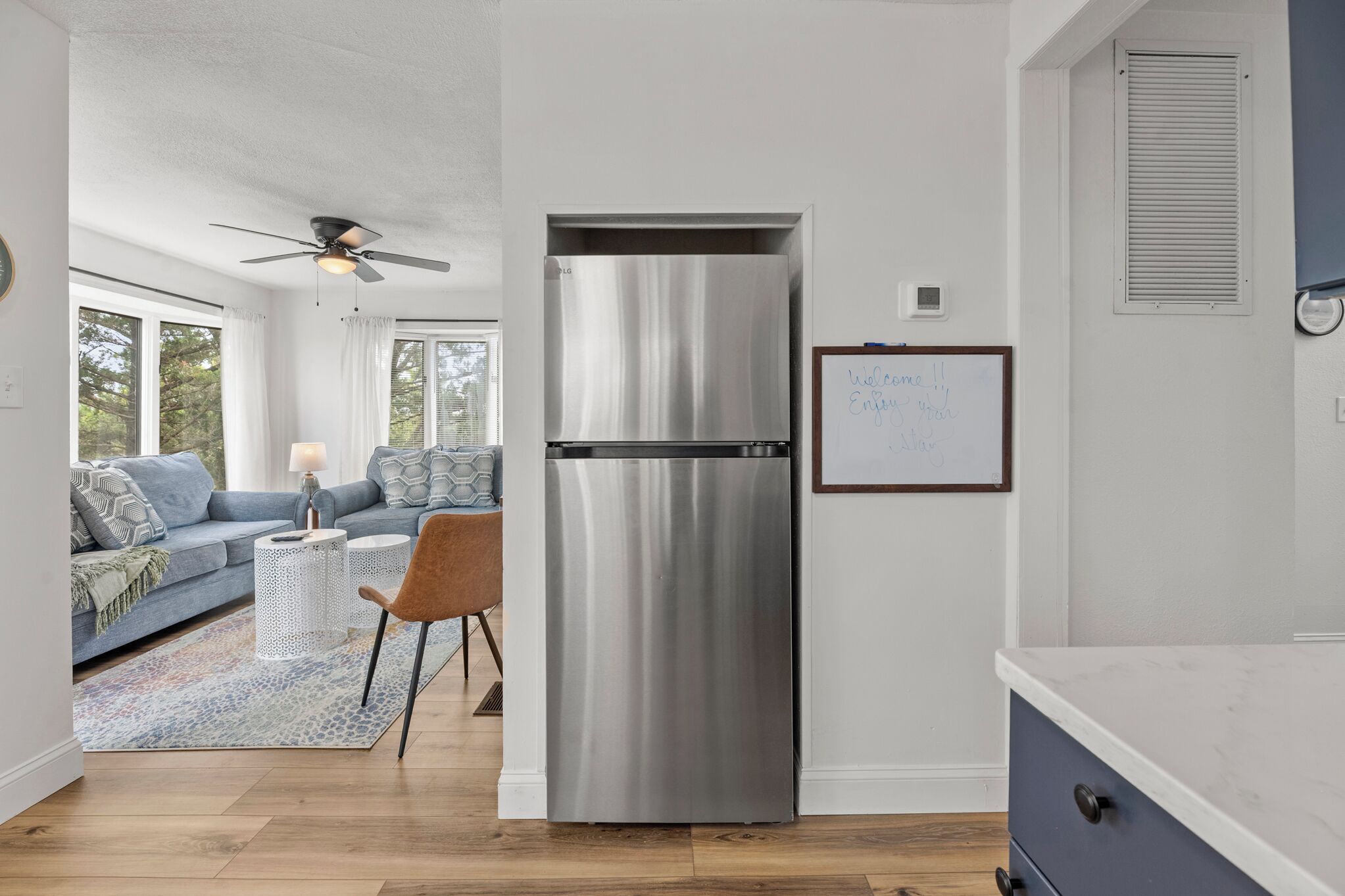 Eine moderne Küche mit einem Edelstahl-Kühlschrank, ein Essbereich mit einem blauen Sofa und einem Couchtisch, sowie ein Blick ins Freie durch große Fenster.