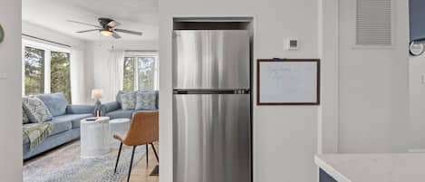 Una cocina moderna con un refrigerador de acero inoxidable, un comedor con un sofá azul y una mesa de centro, y vistas al exterior a través de amplios ventanales.