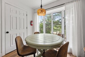 A dining area with a round table, four chairs, a hanging light fixture, a fire extinguisher, and a window with curtains. - Yellow Submarine: Oceanfront | Hot Tub, Deck (Salvo)