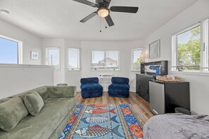Un espacio habitable compacto con ventilador de techo, un sofá verde, dos sillones de peluche azules, una zona de cocina con refrigerador y un tapete estampado.